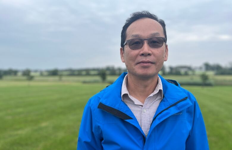 A man with tinted sunglasses and a blue rain jacket stands in an open field.