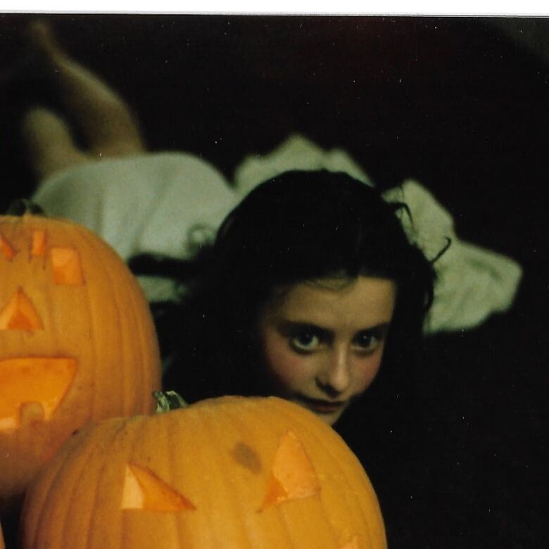 A young child poses next to two jack-o-lanterns on Halloween. 