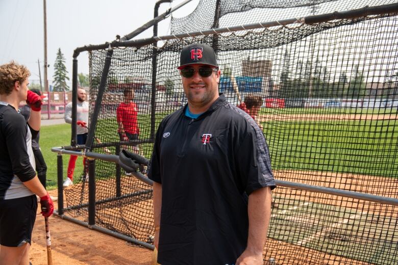 A smiling man wearing sunglasses and Thunder Bay Border Cats clothes stands on a baseball field.