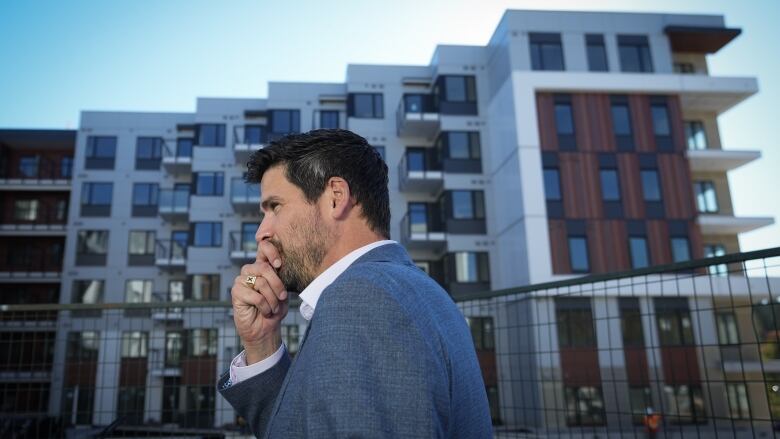 A politician stands to the side while an outdoor announcement in summer is happening. There's a condo-style building in the back.