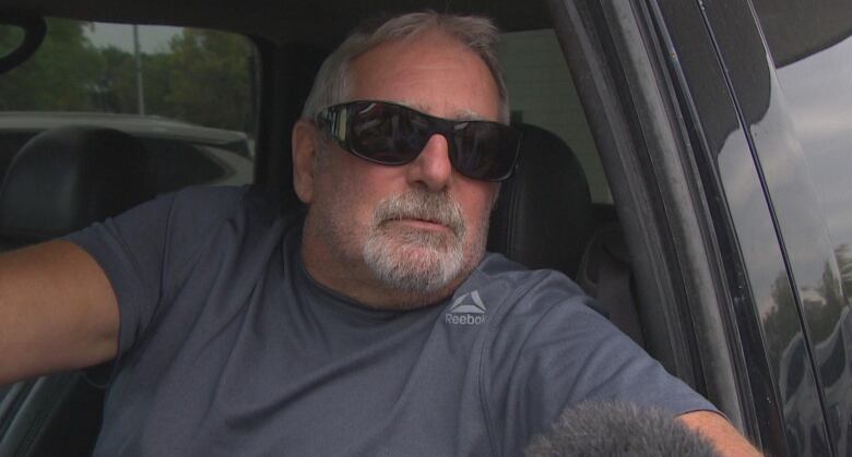 A man wearing sunglasses is shown speaking into a microphone from the driver seat of his vehicle.