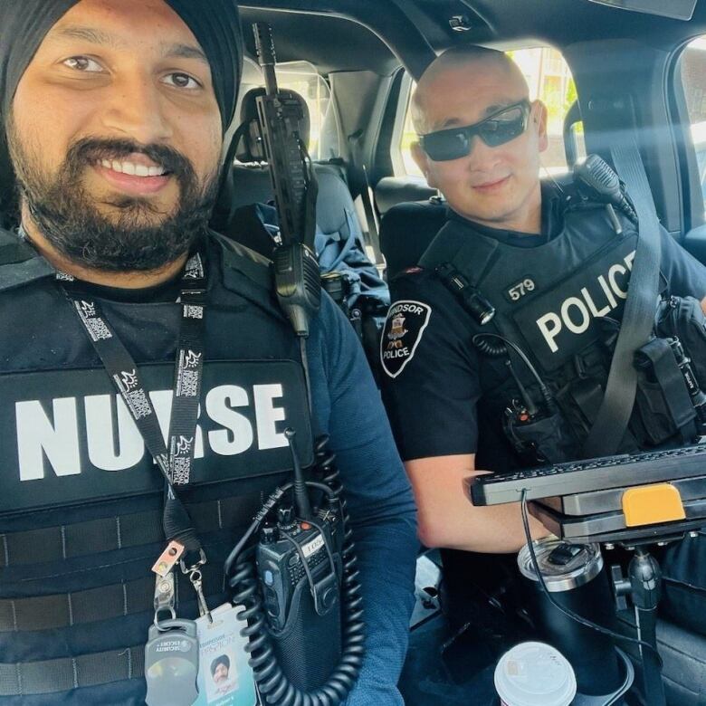 Two people in a police car, one wearing a vest reading 