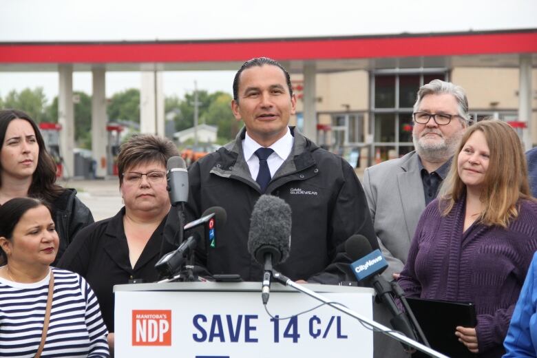 A man in a fall jacket stands at a podium, in front of a gas station across the street.