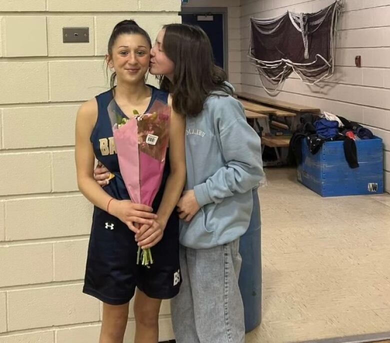 A woman in grey, right, kisses another woman in blue who is holding a bunch of flowers.