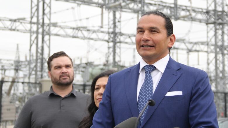 A man speaks at a news conference, standing in front of hydro infrastructure. 