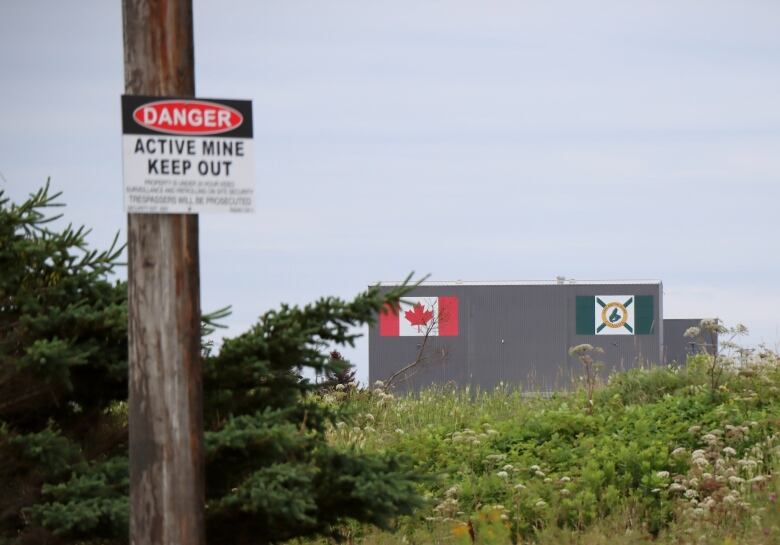 A large grey building with Canada and Cape Breton flags is shown in the distance, with a sign mounted on a pole in the foreground saying 'Danger Active Mine Keep Out.'