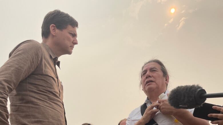 Two men stand together in a dusty field speaking.