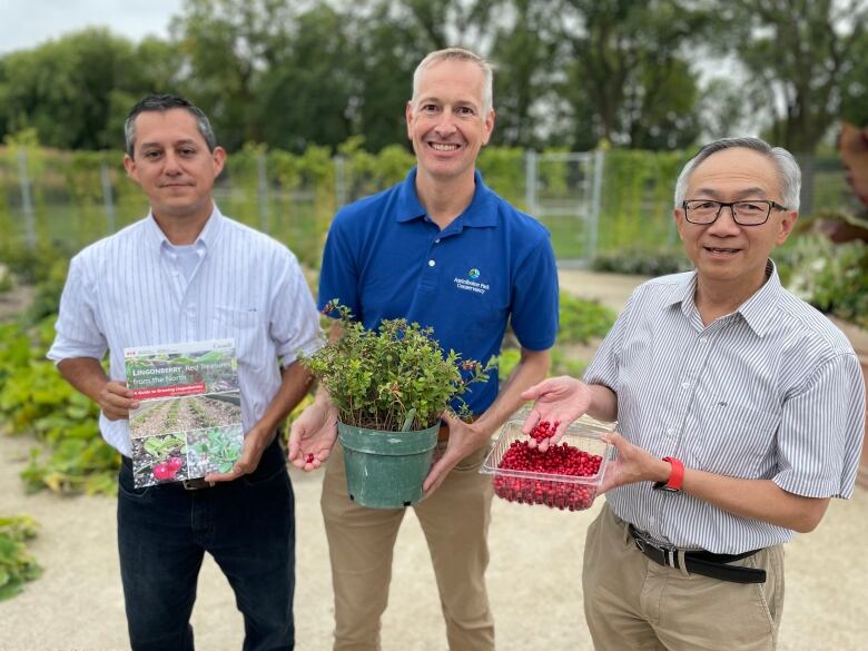 Three men pose for a photo. One holds a container full of red berries, one holds a plant, while the other holds a pamphlet. 