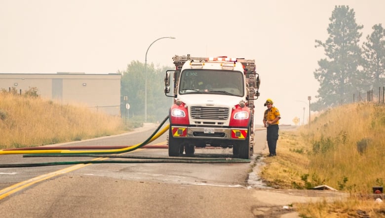 A wide shot of a firefighter standing next to a firefighting vehicle putting out spot fires under a hazy smoky sky.