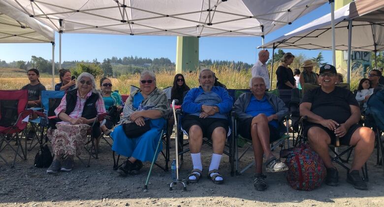 A group of First Nations people look on while sitting under the shade of a tent.