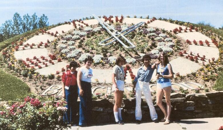 Group of five young people wearing 1970s-style clothing posing in front of a large floral clock.