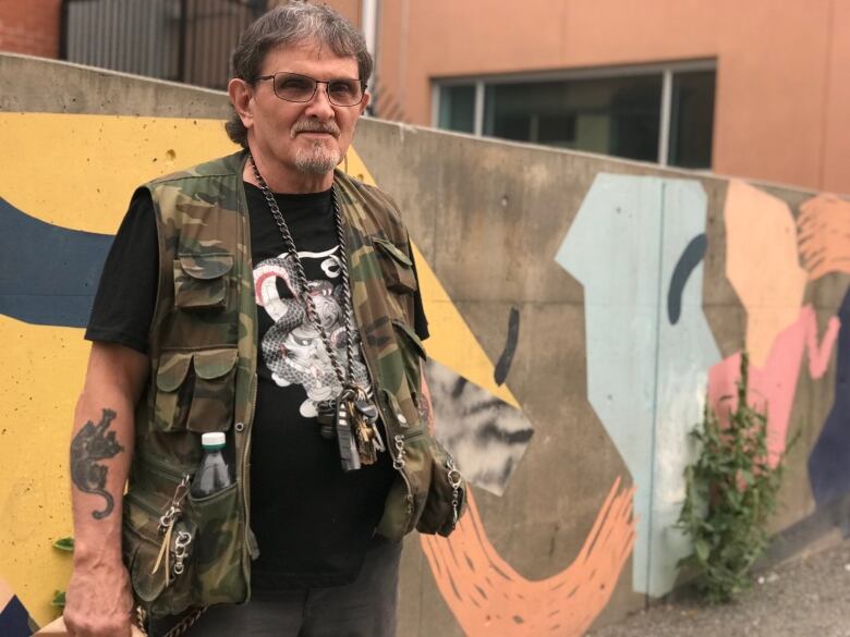 A man with grey hair, glasses and a goatee stands against a wall. He has a camouflaged vest on and tattoos on his arms.