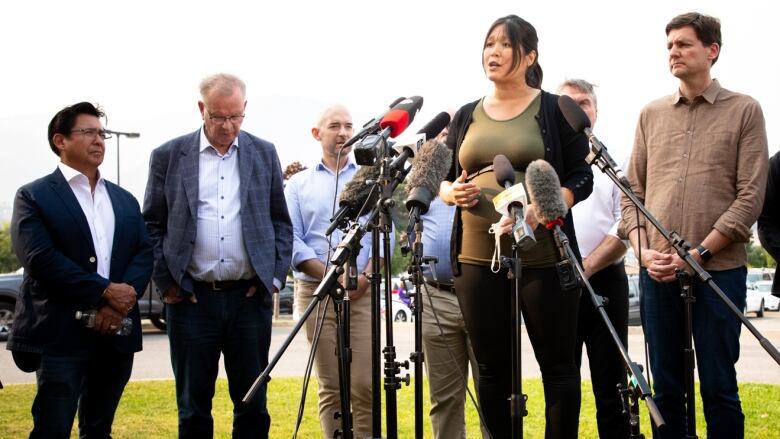 A woman stands on an equipment box to speak to a number of microphones, with a number of men flanking her.