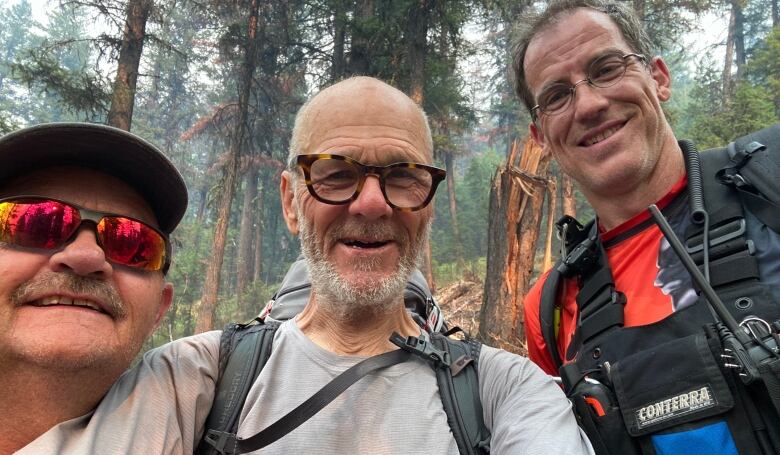 A man in glasses is flanked on either side by a man wearing red sunglasses and a hat and another man with a safety vest and radio  all of them smiling at the camera.