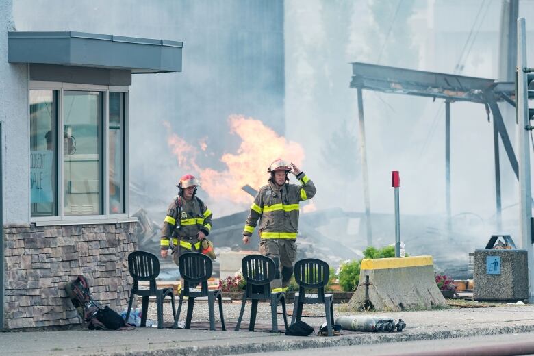 Two firefighters walking with a structure on fire behind them.