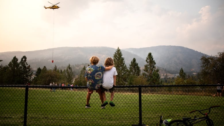 Two children sitting on a wire fence watching a helicopter, with smokey from wildfire and mountain range in the background.