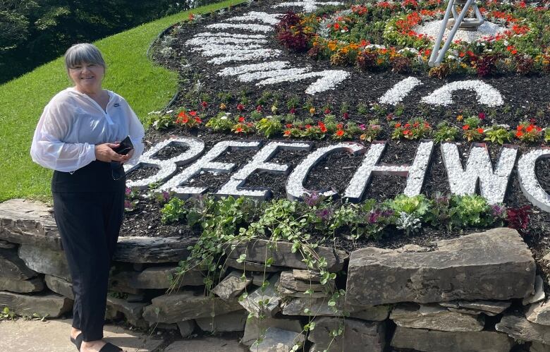 A woman stands in front of a large floral display on the grass that says 