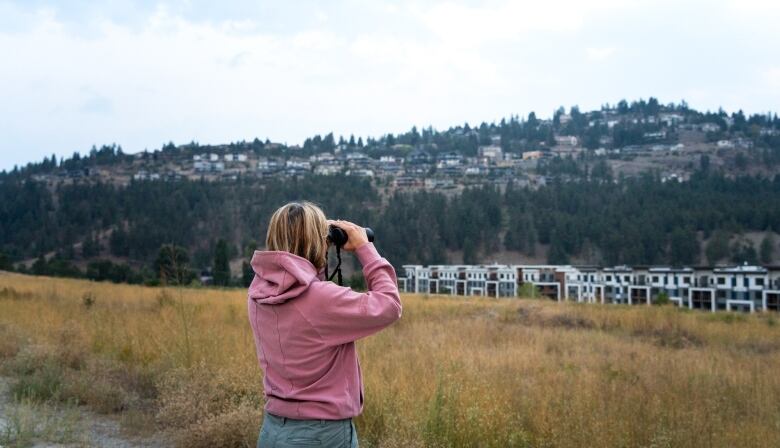 A person with long blonde hair wearing a pink hoodie looks through binoculars at the hillside dotted with homes.