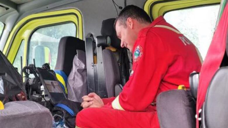 A man in a red firefighter suit, hands clasped, sits in a vehicle looking at a screen.