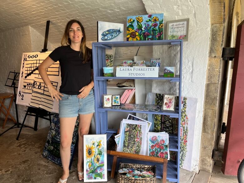 Woman in black t-shirt and denim shorts stands beside a blue bookshelf of floral and landscape paintings. 