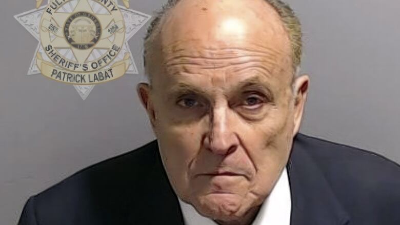 Balding man in suit with neutral face stands beside Sheriff's Office emblem