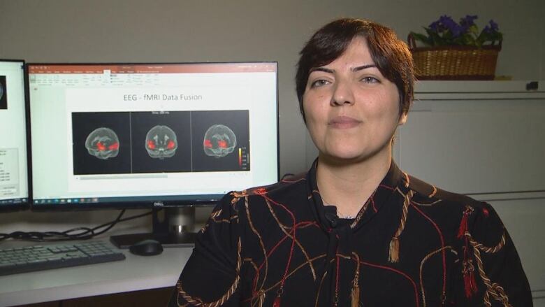 A woman sits in front of a computer screen that has images of a brain behind her.