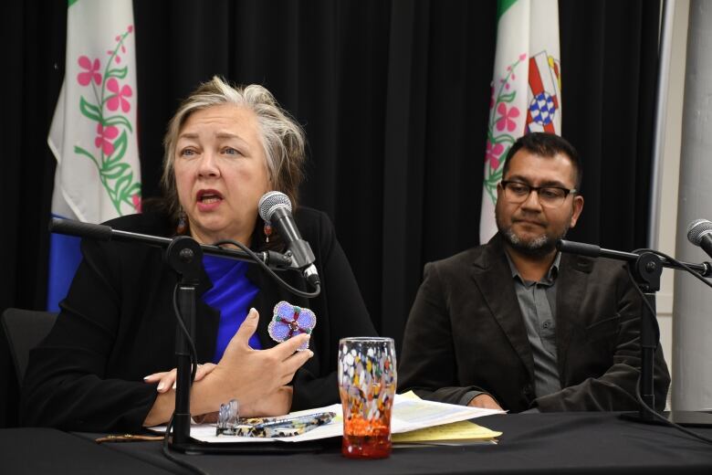 A woman sits at a table speaking into a microphone, while a man sits beside her, looking on.