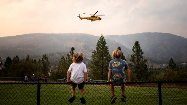 Two children sit on a chainlink fence, watching a yellow firefighting helicopter hover in the air.