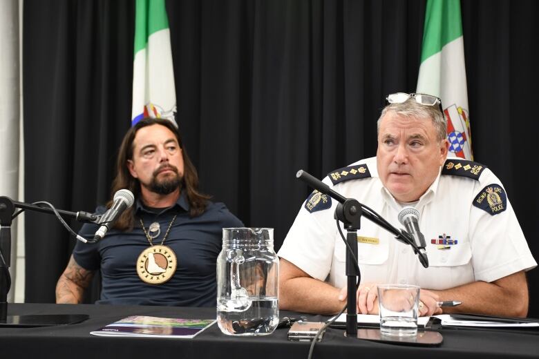 A man in an RCMP uniform shirt sits at a table speaking into a microphone, while another man sitting beside him looks on.