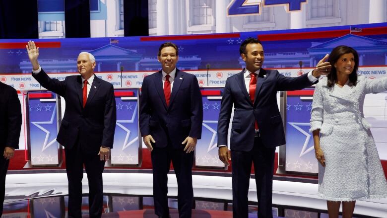 3 men in dark suits and a red tie and a woman in a light coloured dress on a stage, waving and smiling