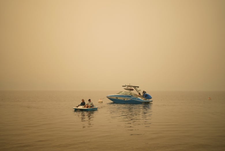 A larger boat is seen in the background of a paddleboat being driven by two people, with wildfire smoke hanging in the air.