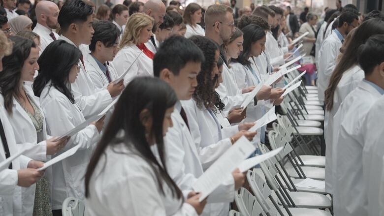 A group of people in white coats are pictured lined up in rows, looking down at a piece of paper in their hands.