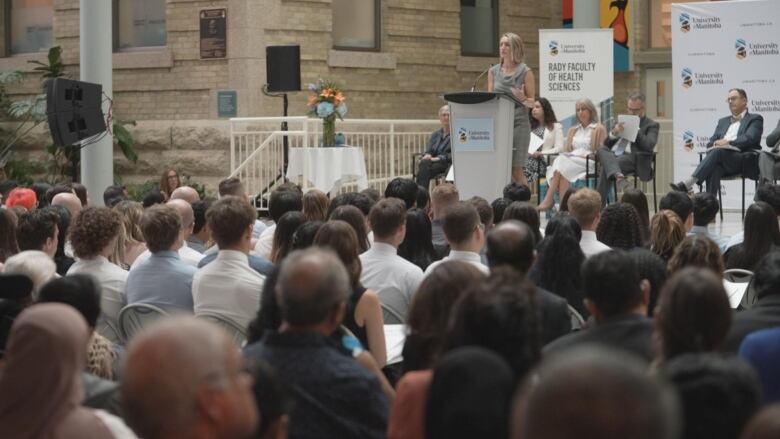 A woman is shown speaking at a podium to a large group of people.