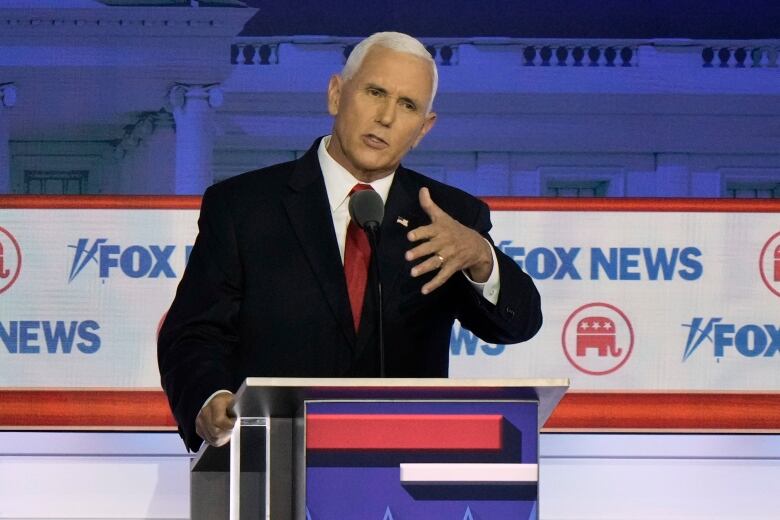 White haired man gestures as he speaks from behind a podium