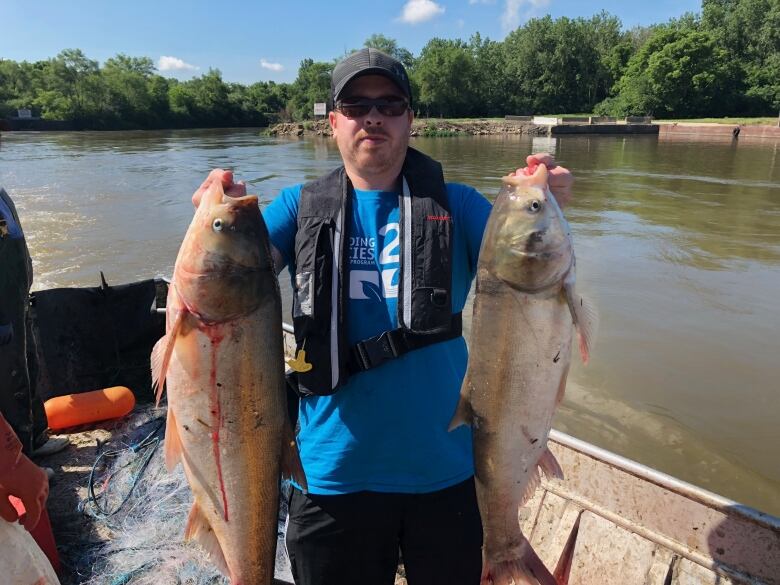 A man in a baseball cap, standing on a boat, holding up two large fish. 