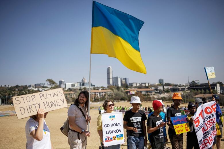 Several people hold signs in an outdoor demonstration. One reads, 'Stop Putin's Dirty Money,' while another says, 'Go Home Lavrov.' Another person in the small group holds a large Ukrainian flag.