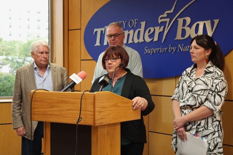 Four people stand around a podium while one woman speaks into a microphone.