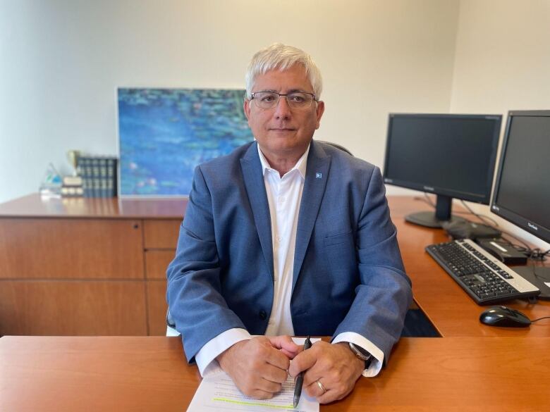 A man with white hair, wearing a blue suit, sits at his desk.