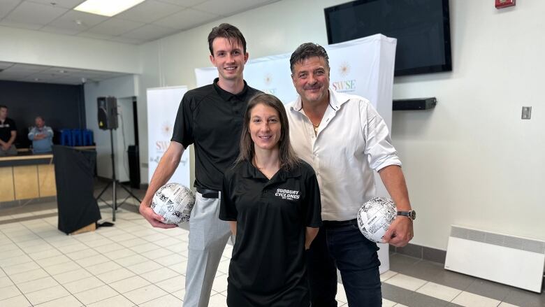 A tall man in a black t-shirt holding a football in his left hand, a man in a white shirt holding a football in his right hand. a woman in a black t-shirt that says Sudbury Cyclones standing in front of them.