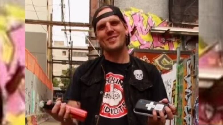 A man in a dark jacket over a black shirt with a round red emblem that features a hand giving the finger smiles at the camera, holding a naloxone kit in one hand and pepper spray in the other.