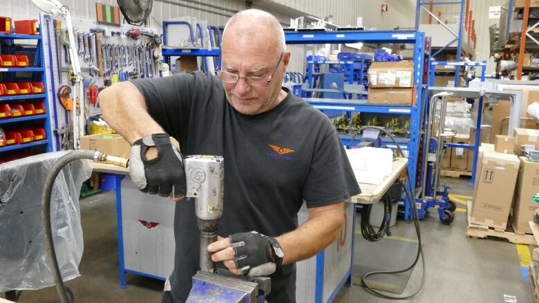 A man stands at a tool bench holding a pneumatic tool, bolting something onto a piece of equipment held in a vice