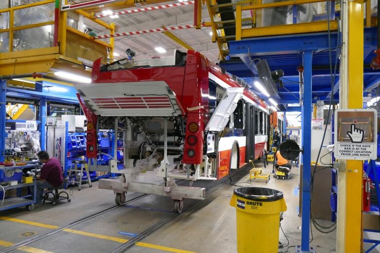 A large, partially built red and white bus stand on New Flyer's assembly line, surrounded by yellow ladders and other equipment.