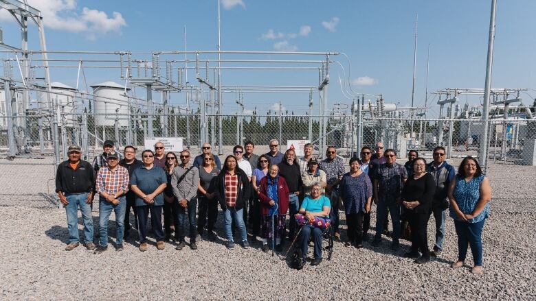 A large group of people standing in front of a chain-link fence, behind which is a field of gray electrical infrastructure.