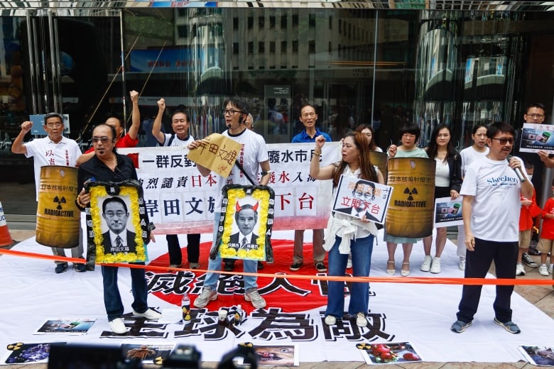 More than a dozen protesters, some with their fists raised, hold up placards featuring images of men and signs in Japanese while one of them speaks through a microphone.