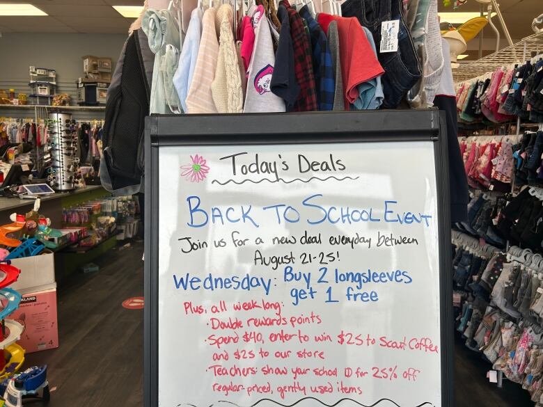 A whiteboard placed in front of a rack of clothing at Once Upon A Child in Winnipeg advertises deals on gently used kids clothing. It reads 