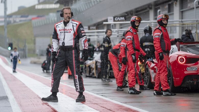 A man wearing race gear steps out onto a racetrack. Behind him are race cars and race crews.