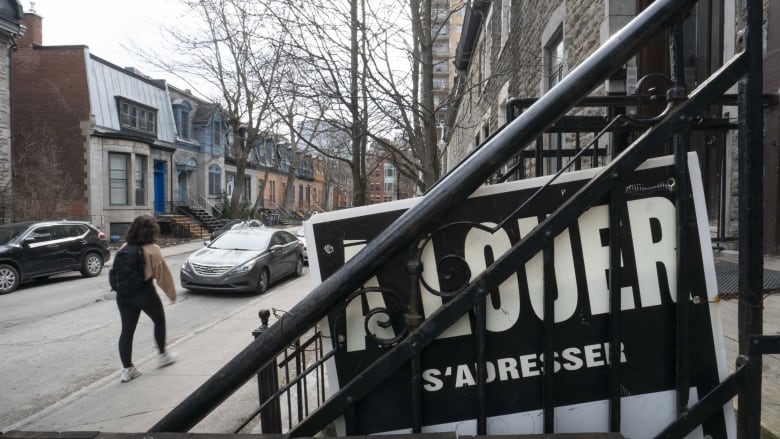 A sign that says for rent in French, on a staircase railing. A young woman walks on the street nearby.