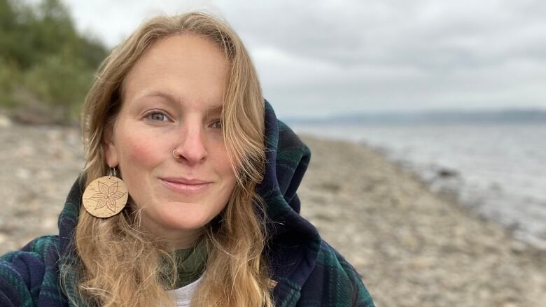 A woman with long hair and a nose ring takes a selfie of herself standing near shore.