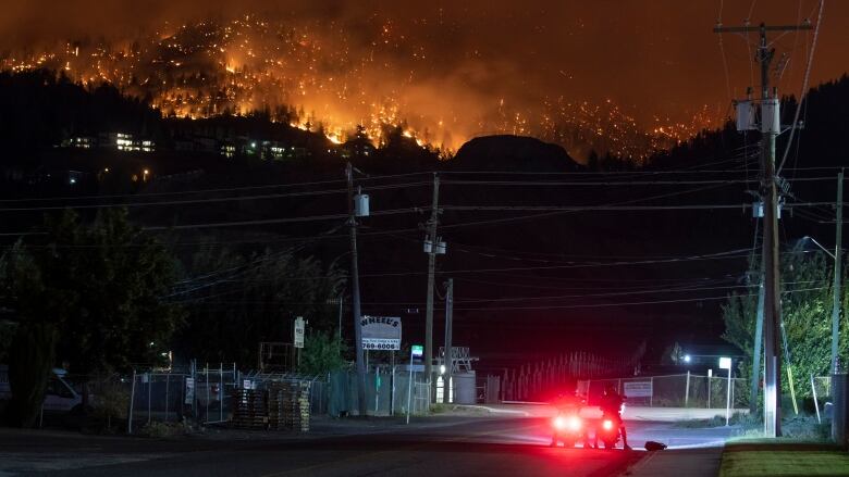 Two motorcyclists stop at an intersection and watch a wildfire burning on a nearby mountain ridge.