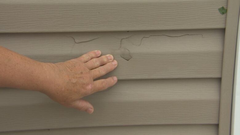 A person's hand is seen close-up, pointing out a broken section of siding on a garage